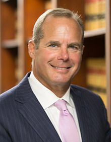 Man in a tan suit and glasses, hands clasped, looking at the camera. He is in an office with a window behind him.