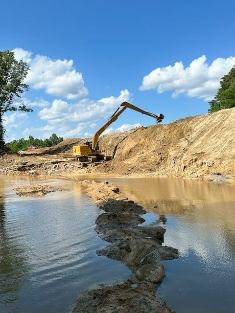 Excavator removing dirt near a body of water under a partly cloudy blue sky.