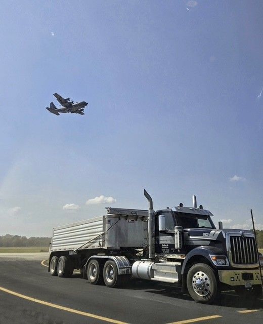 Semi-truck on a road with a large airplane flying in a clear blue sky.