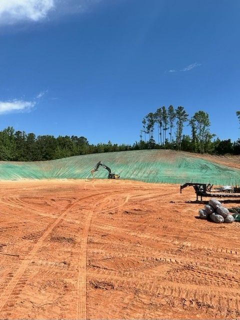 Construction site with an excavator working on a hillside covered in green netting under a blue sky.