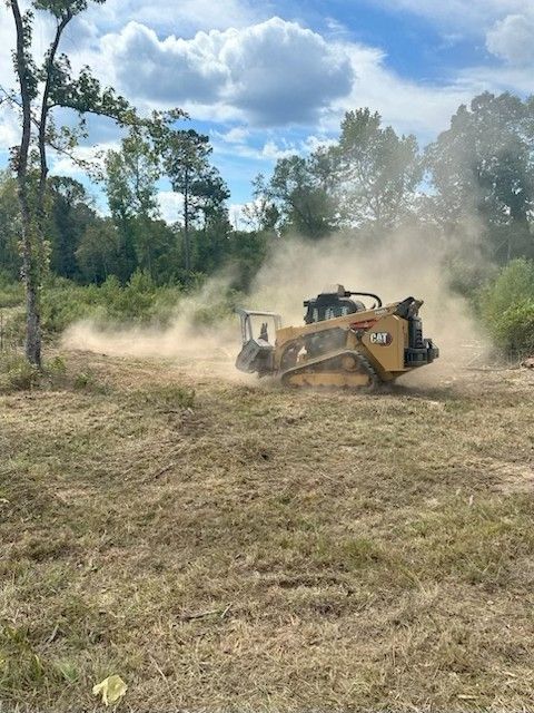 Yellow Caterpillar machine clearing brush in a field, kicking up dust under a cloudy sky.