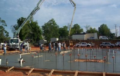 Construction site: Concrete being poured from trucks into a wooden-framed foundation. Workers supervise on a sunny day.
