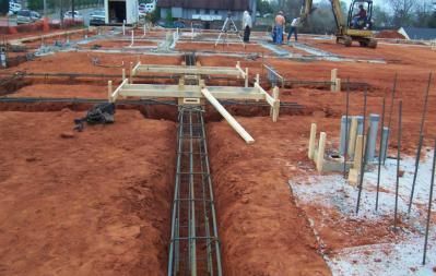 Construction site with foundation framework and rebar, workers, excavator, and red soil.