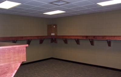 Empty corner room with long wooden shelf supported by brackets. Beige walls, brown trim, and carpet.