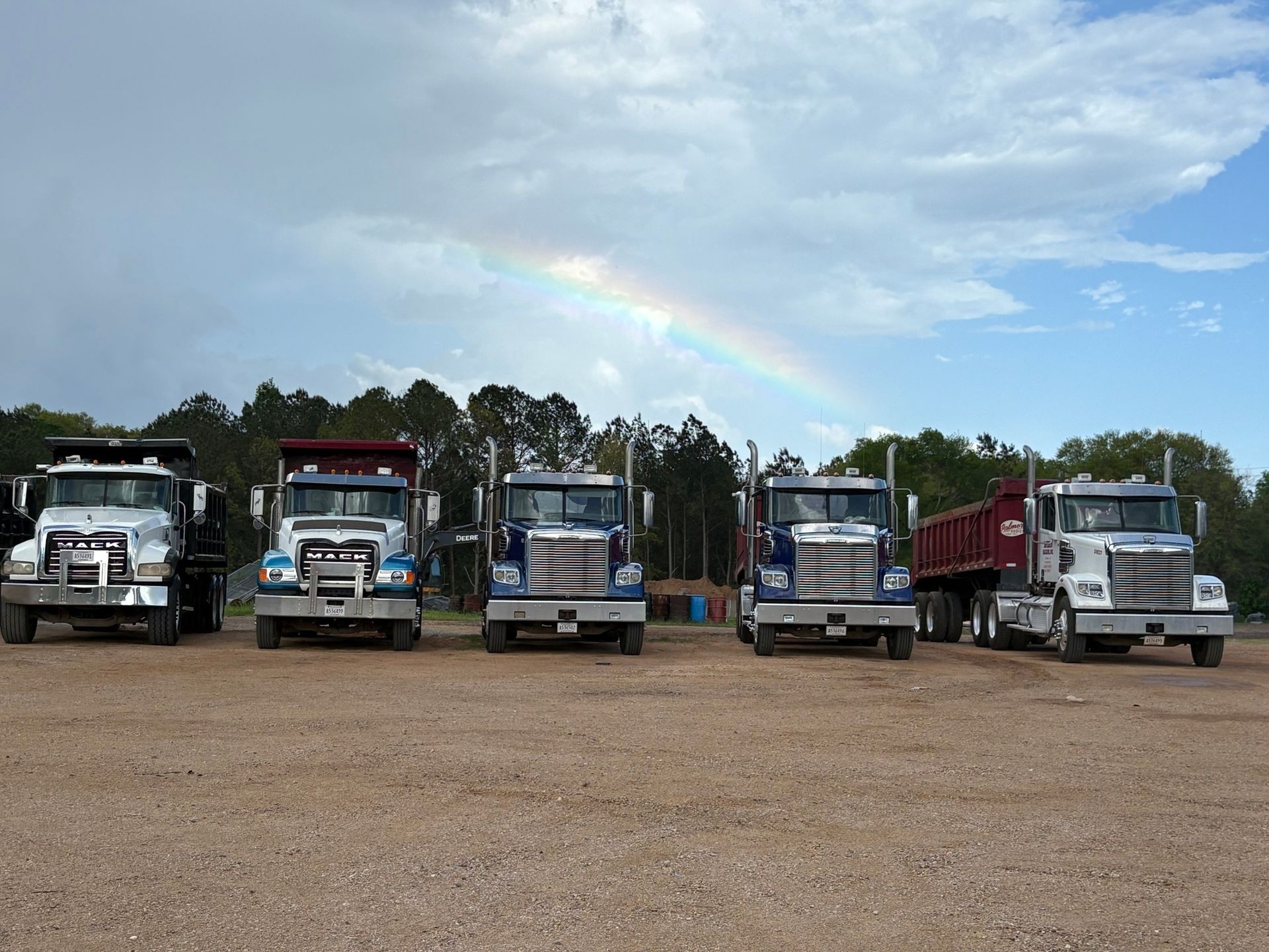 Five dump trucks parked outdoors with a rainbow in the background.