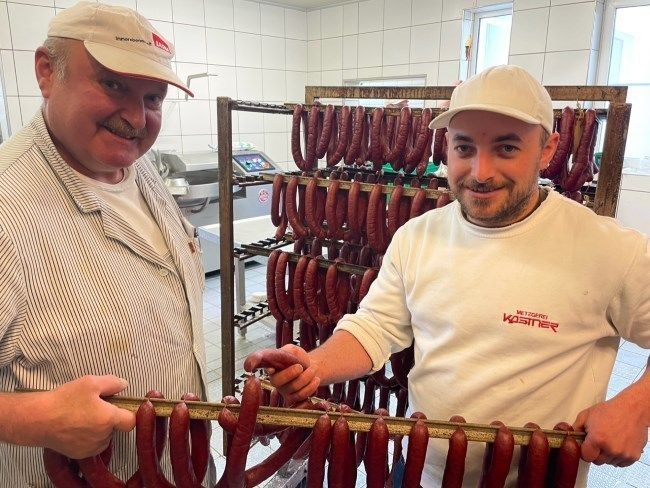 Zwei Männer in einer Werkstatt halten aneinandergereihte Würste auf Metallgestellen.