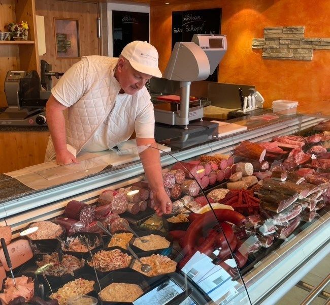 Ein Ladenbesitzer arrangiert Fleisch und Käse in einer Feinkosttheke unter warmem Licht.
