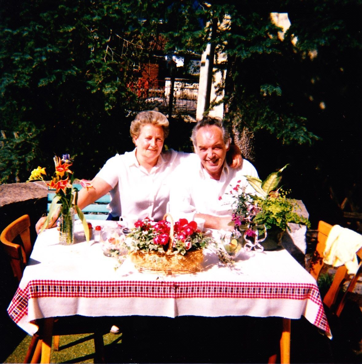 Zwei Personen sitzen an einem mit Blumen geschmückten Gartentisch und lächeln in die Kamera.