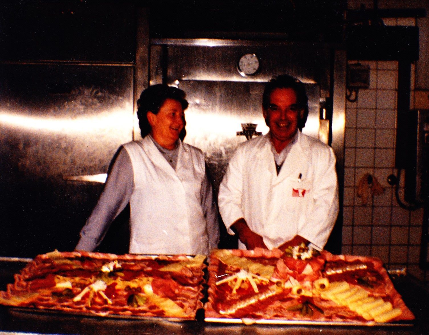 Zwei Köche in weißen Kitteln stehen hinter Tabletts mit Pizza in einer Küche.