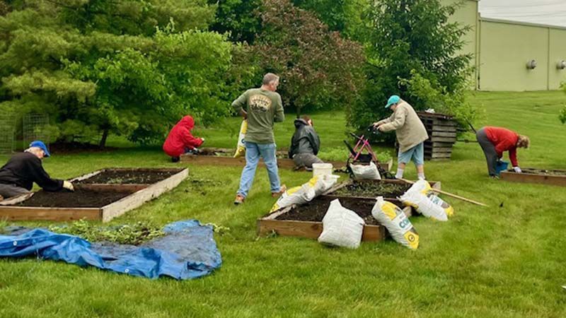 People gardening in raised beds outdoors, filling them with soil. Bags of soil sit nearby.