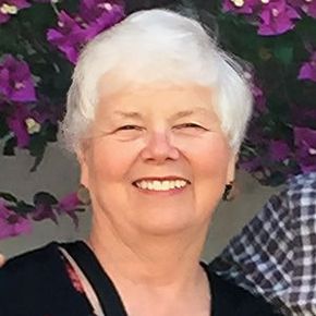 Woman with white hair smiling in front of purple flowers, wearing black shirt.