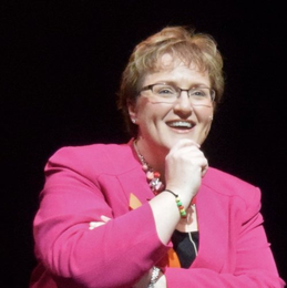 Woman in pink jacket, wearing glasses, smiling and gesturing, on stage with dark background.
