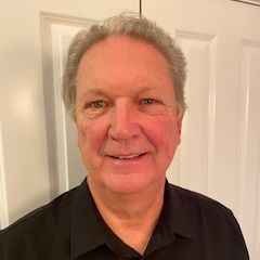Man with gray hair smiling, wearing a black shirt, in front of a white door.