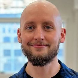 Bald man with a beard, smiling, wearing a blue plaid shirt and black shirt, in front of a window.