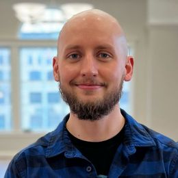 Bald man with a beard, smiling, wearing a blue plaid shirt and black shirt, in front of a window.