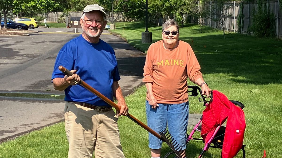 Two people raking grass on a lawn. One holds the rake; the other leans on a walker. Sunny day.