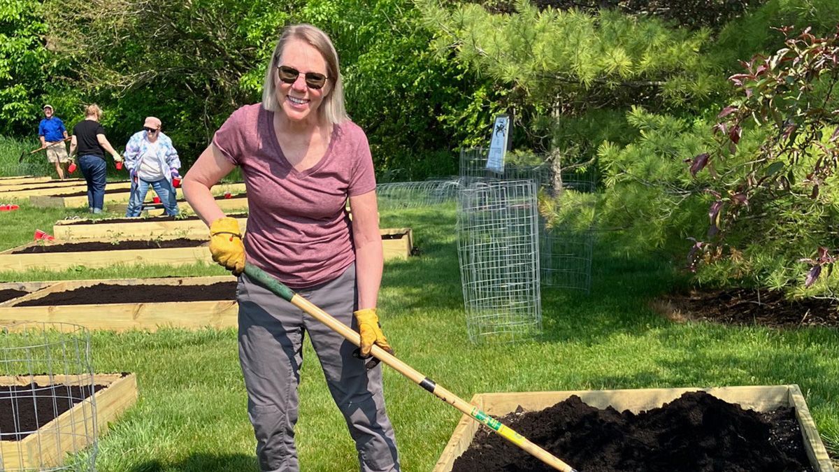 Woman raking soil in raised garden bed; others working nearby on a sunny day.