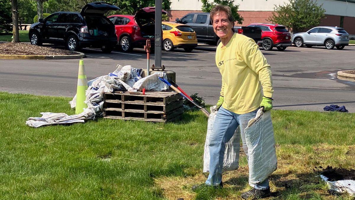Person in yellow shirt carrying debris on a lawn; cars parked in the background.