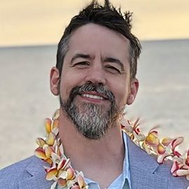 Man wearing a flower lei, smiling at the camera, with a graying goatee.