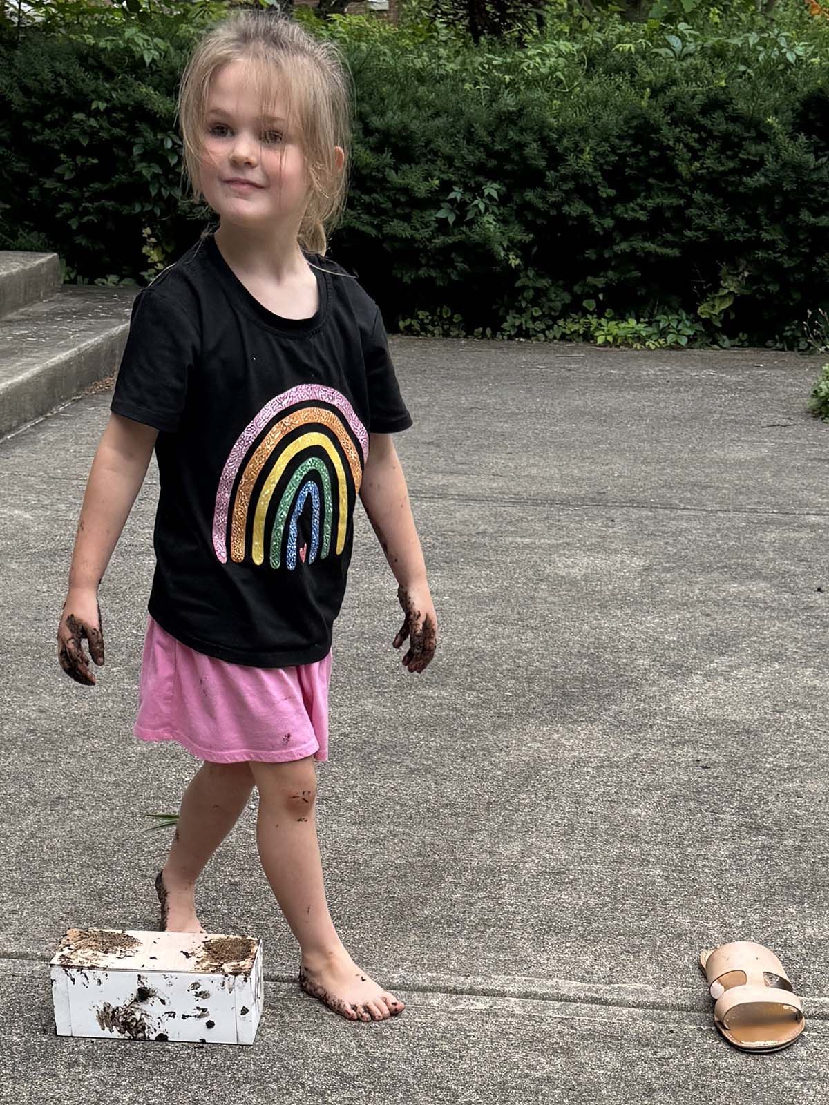Child standing on a driveway in a black rainbow T-shirt and pink shorts, near a box and sandals.