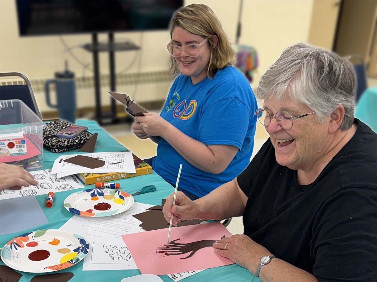 Two women painting at a craft table with palettes, brushes, and papers, smiling in a classroom setting