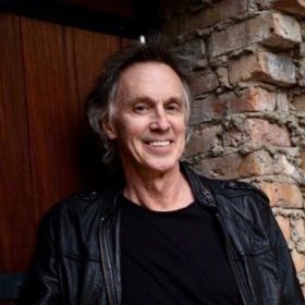 Man smiles in black leather jacket, leaning against brick wall, doorway at left.
