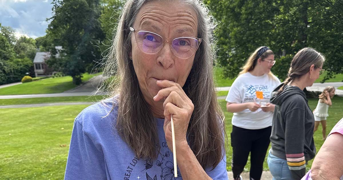 Woman with purple glasses stands outdoors on a grassy lawn, with people in the background.