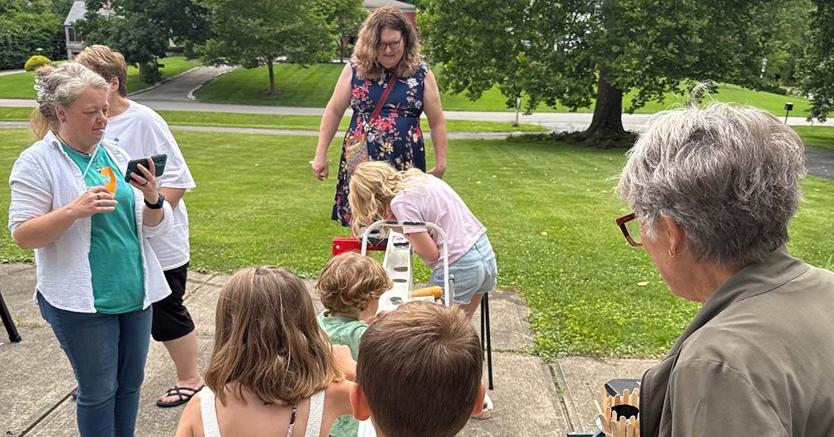 Children and adults gathering outdoors on a park path, with one child reaching into a red bucket