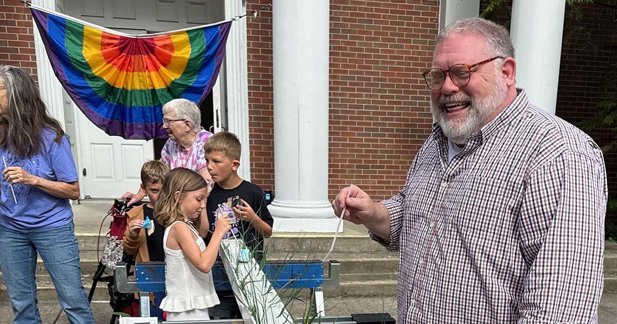 People chatting outside a building, with a rainbow banner and several children gathered nearby.