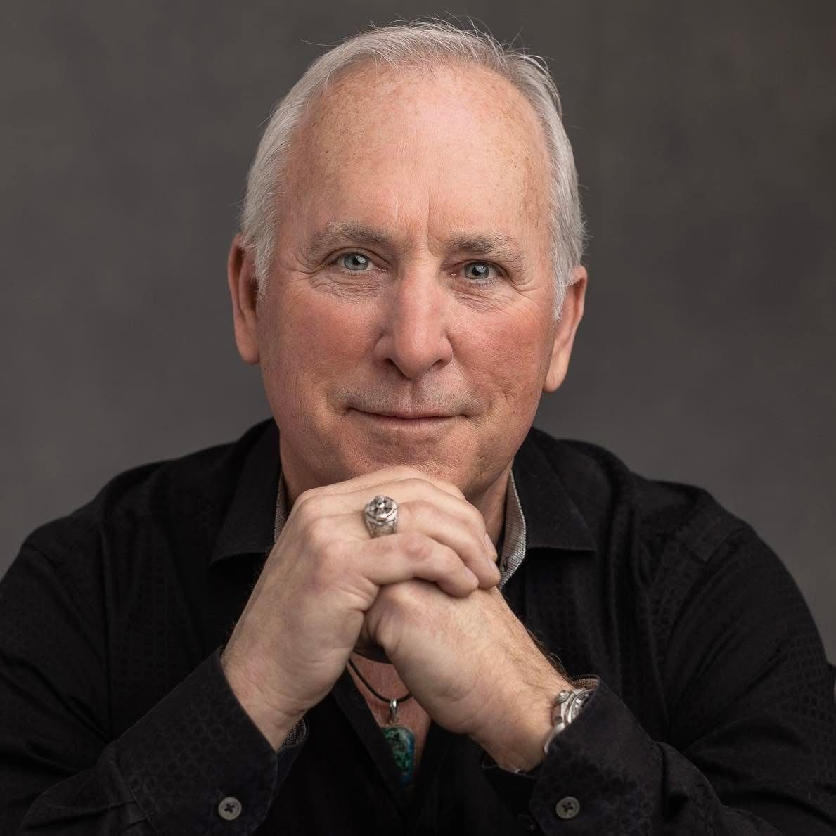 Man with short white hair, resting chin on clasped hands, wearing black shirt and ring.