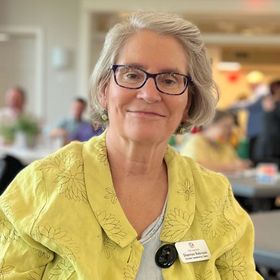 Woman with glasses in a yellow jacket smiles, seated indoors, name tag visible.