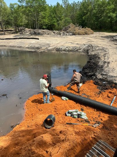 Workers laying orange absorbent booms beside a muddy pond in a wooded area