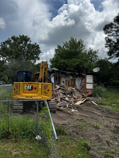 Yellow excavator demolishing a small, damaged house amid debris under cloudy skies