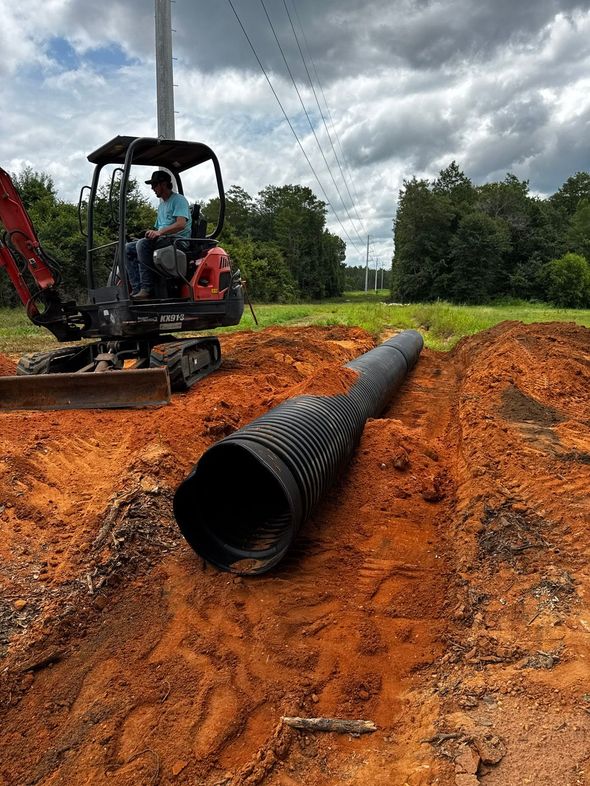 Construction crew laying a large black drainage pipe in a muddy orange trench under cloudy skies