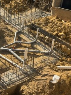 Concrete foundation under construction with rebar, pipes, and excavated soil.