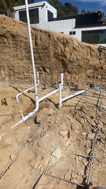 White PVC pipes and fittings on a dirt lot beneath a tan hillside, with houses in the background