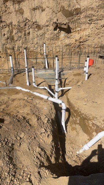 White PVC pipes rise from a dirt trench at a construction site against a bare soil wall.