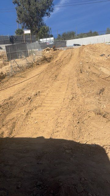 Dirt road or slope under construction, with tire tracks, fence, and trees under a clear blue sky