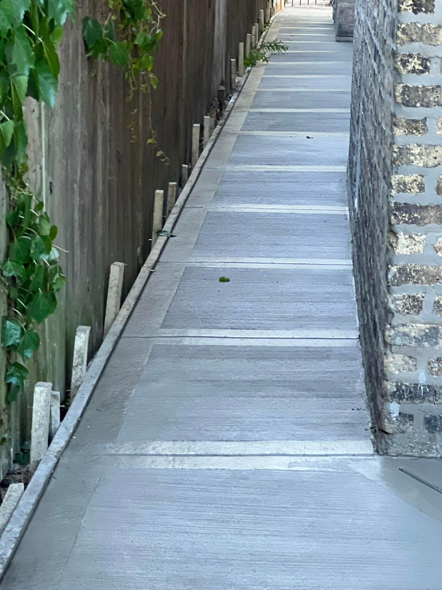 Narrow concrete walkway between a wooden fence and stone wall, with steps ahead.