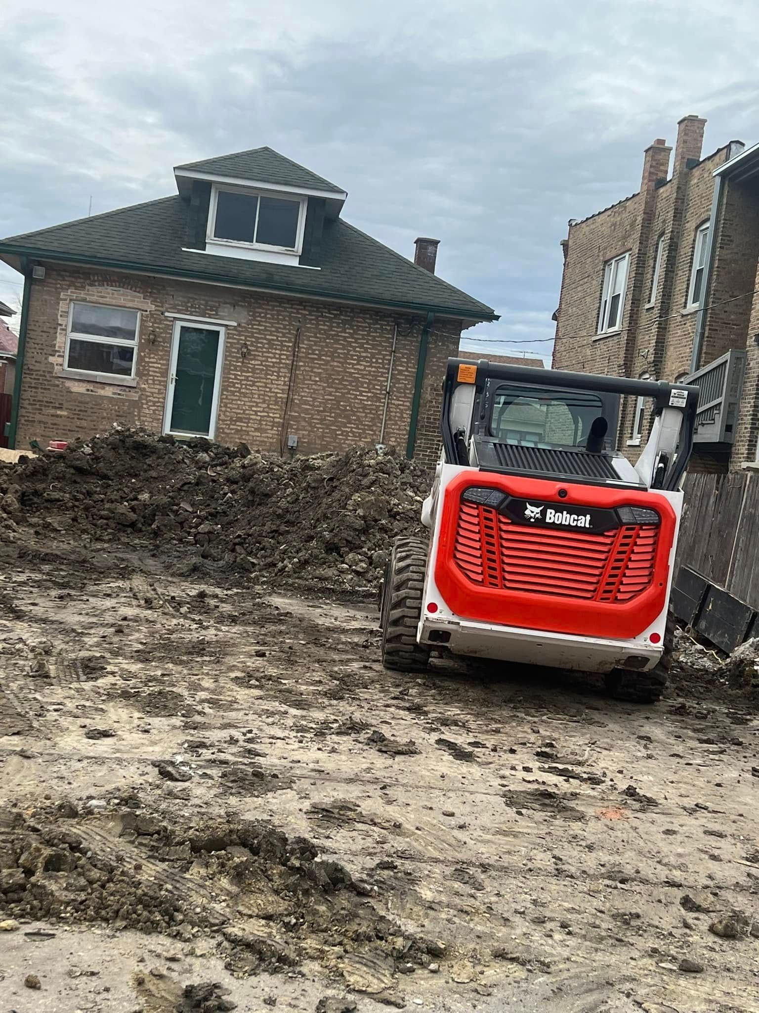 Red Bobcat skid steer in muddy construction site beside a tilted brick house under renovation