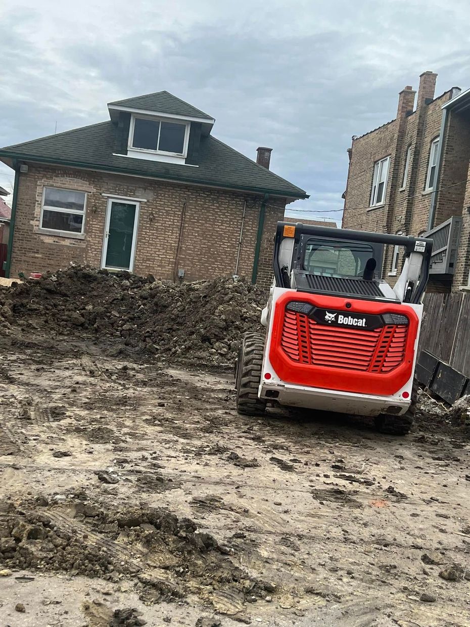 Red Bobcat skid steer in muddy construction site beside a tilted brick house under renovation