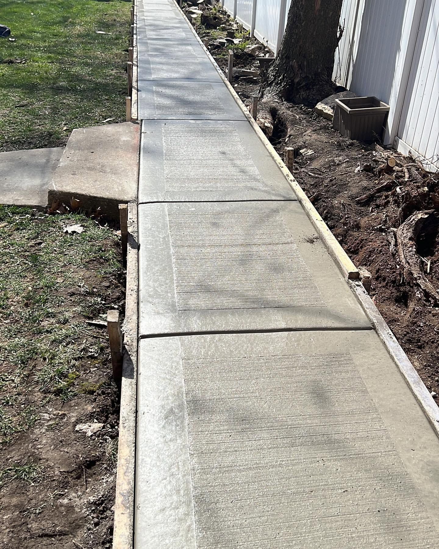 Narrow concrete sidewalk beside a fence, with a curb and grass on one side and dirt on the other.