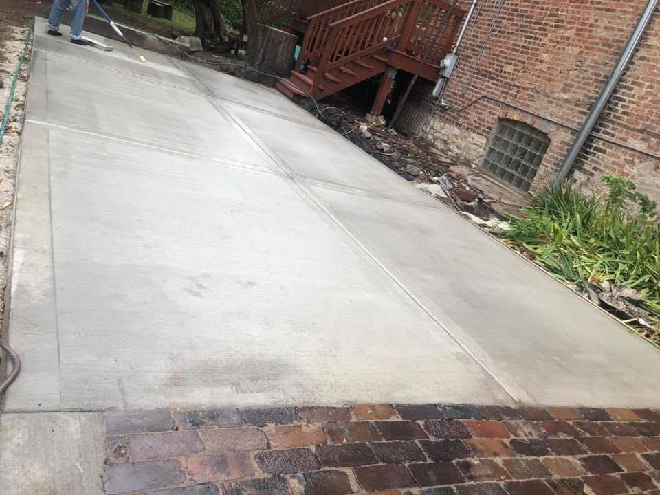 Concrete driveway leading to a brick patio beside a house, with a small wooden deck and plants nearby.