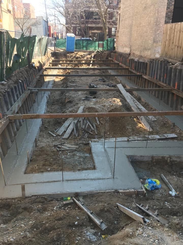 Excavation site with concrete foundation walls, wooden supports, and dirt, surrounded by buildings.