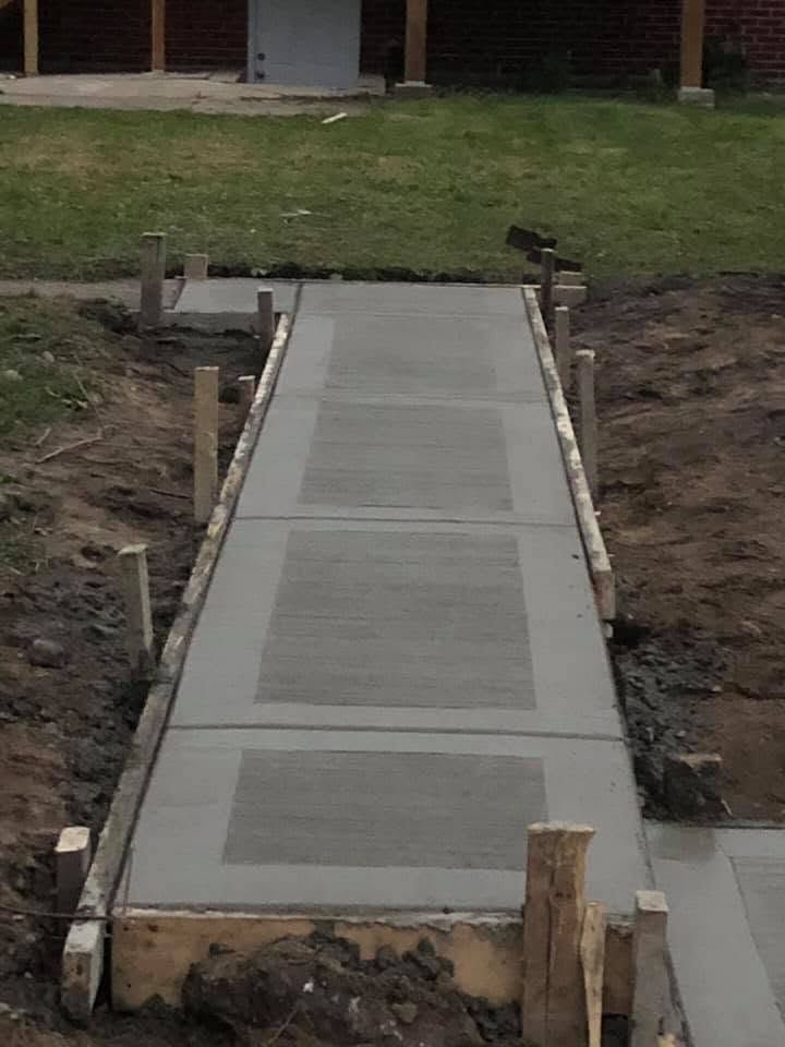 Concrete accessibility ramp under construction leading to a building entrance, with dirt sides and no handrails.