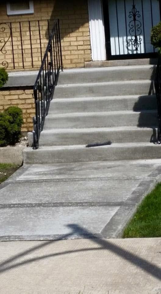Concrete front steps leading to a house entry with black railing and a sidewalk in the foreground