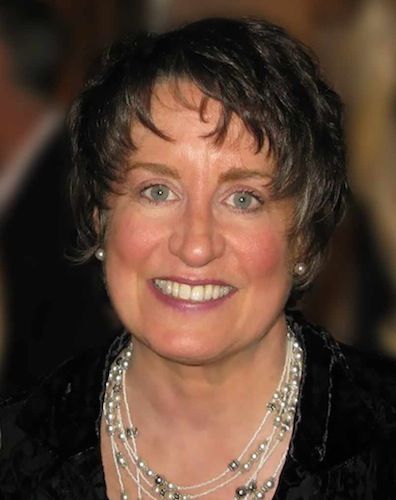 Smiling person with short dark hair wearing a black top and layered silver necklaces against a blurred background