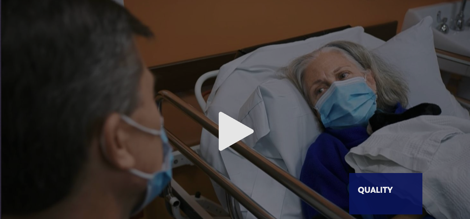Masked visitor speaks to a patient resting in a hospital bed, with a play icon overlay.