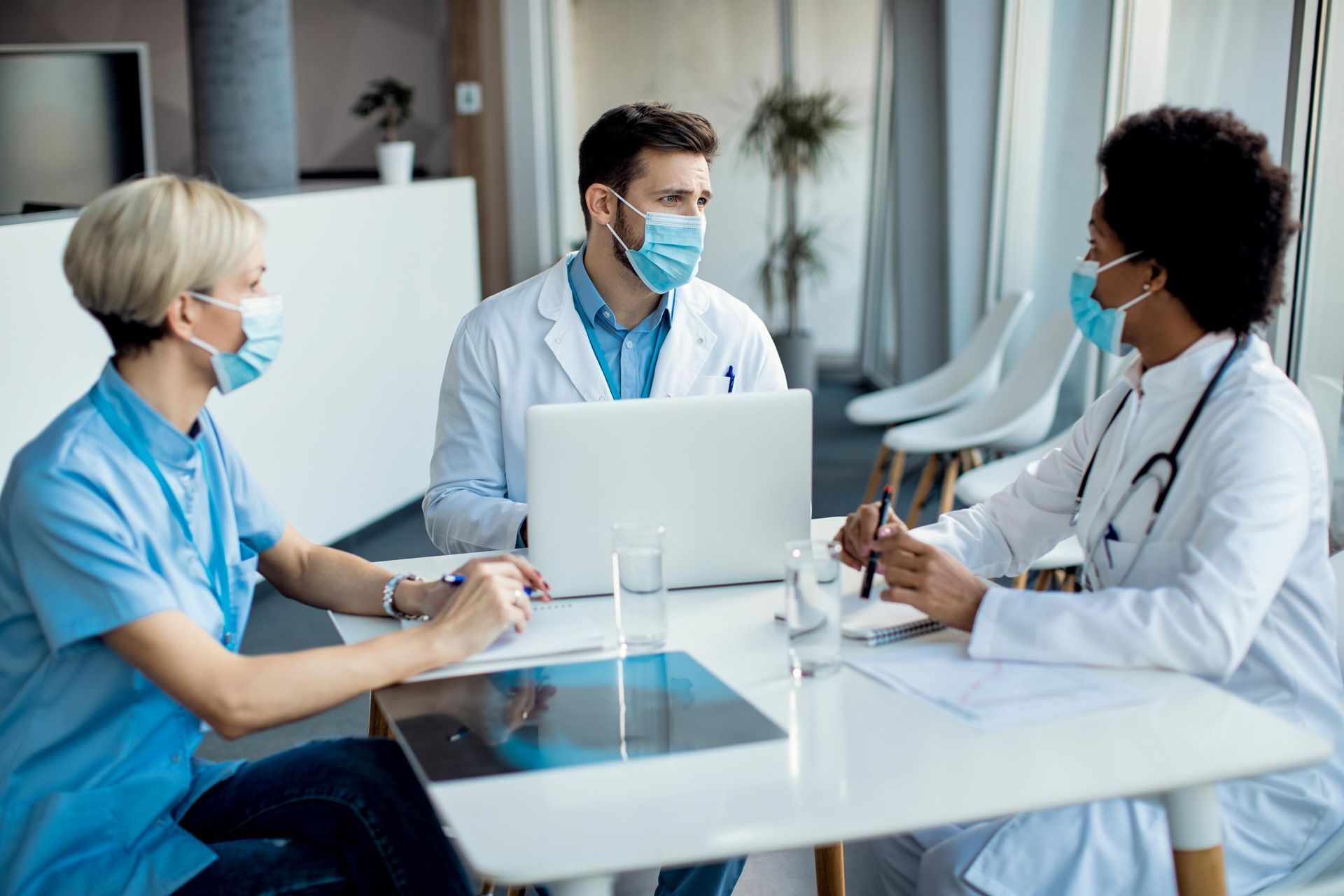 Masked medical staff in a clinic meeting around a laptop at a white table.