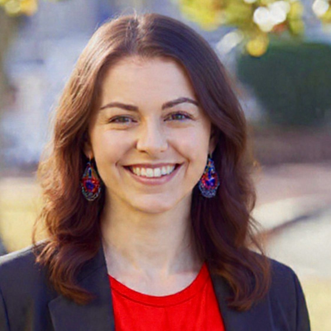 Smiling woman with brown hair and colorful earrings outdoors in a dark blazer and red top